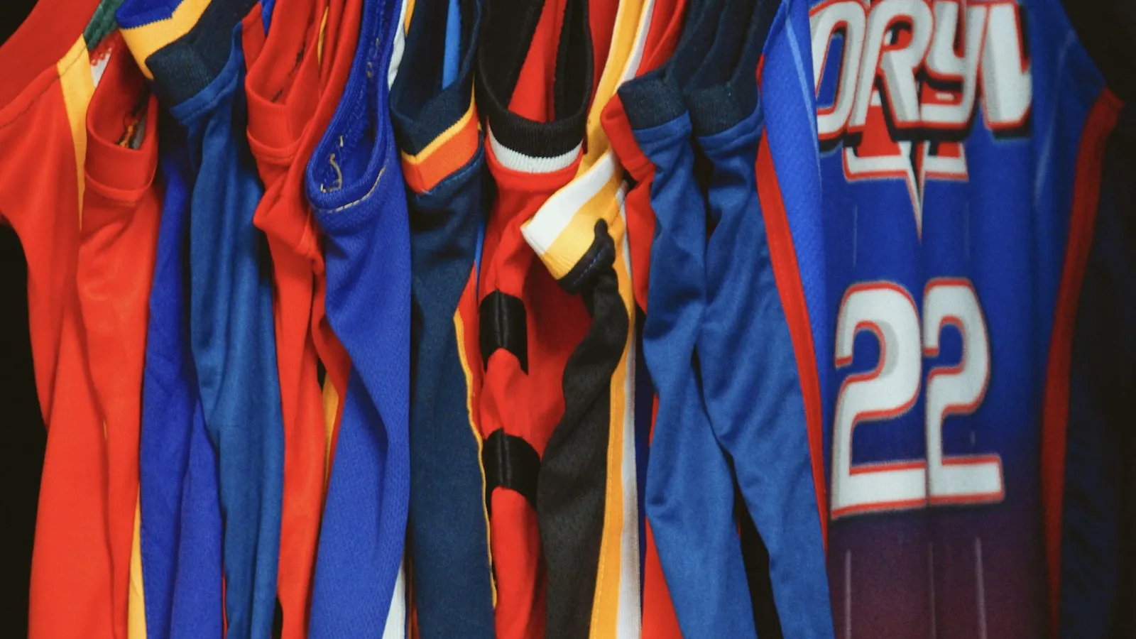 Maillots de basketball numérotés rouge et bleu accrochés sur cintres dans un vestiaire.