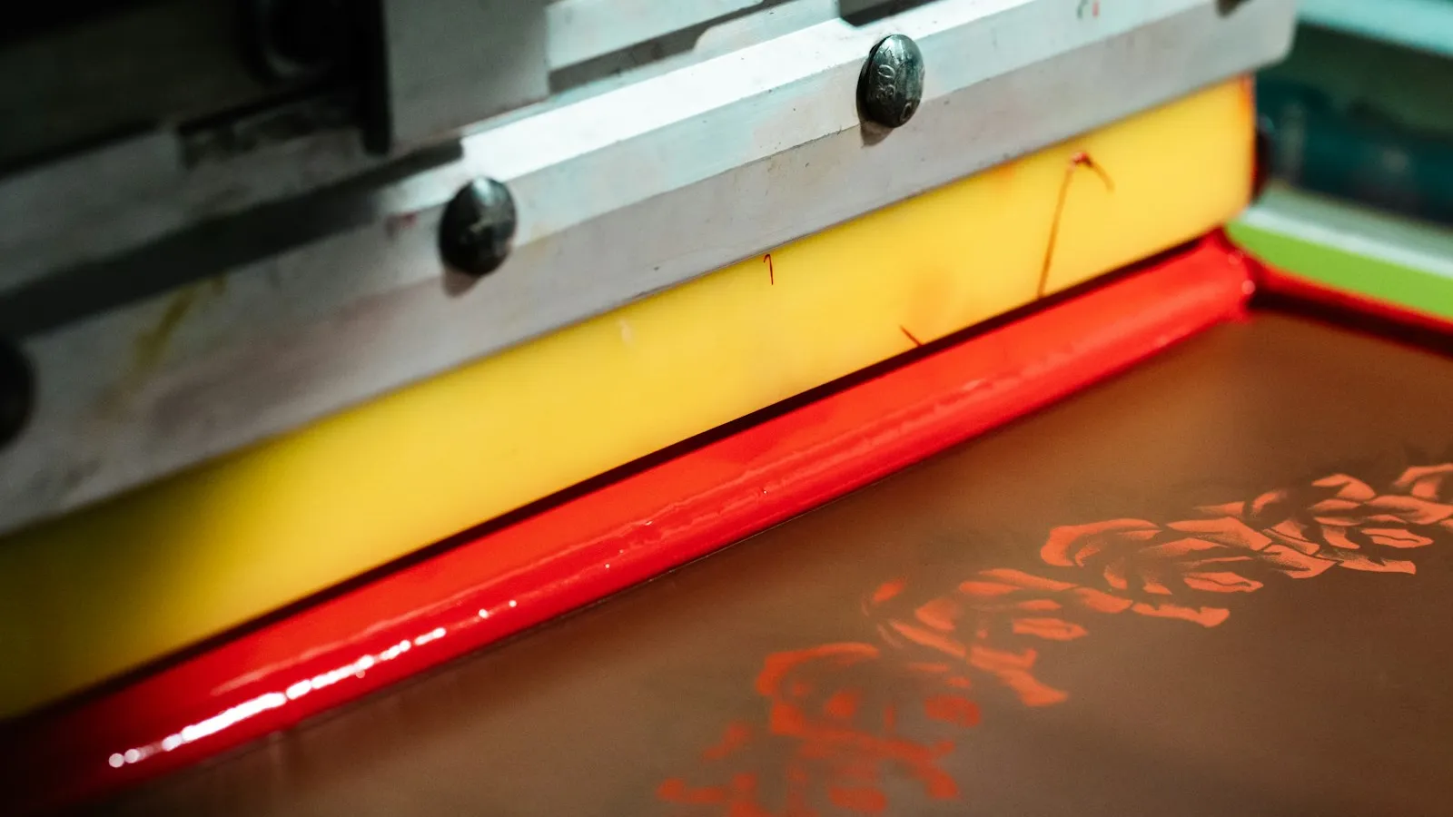 Détail macro d'une racle laissant apparaître une bande d'encre rouge éclatante au-dessus d'un écran de sérigraphie.